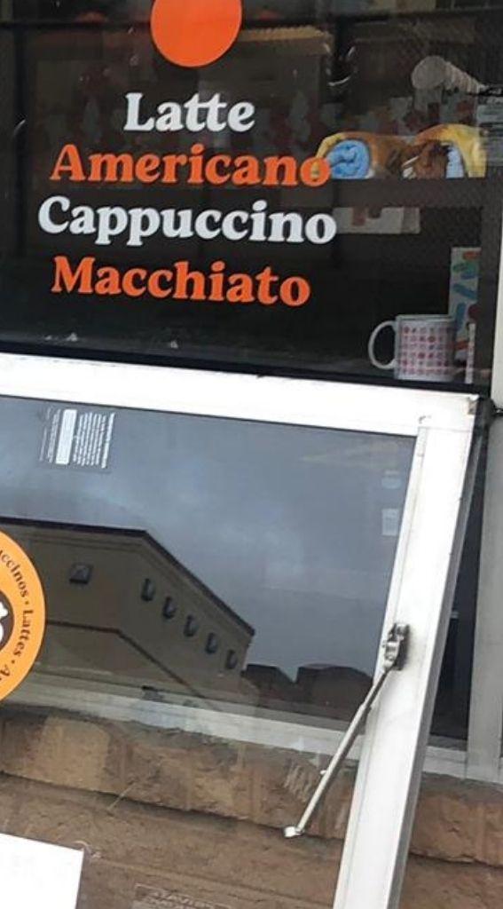 Window display featuring menu items: Latte, Americano, Cappuccino, Macchiato, with a visible orange circle logo, promoting coffee options at a food establishment.