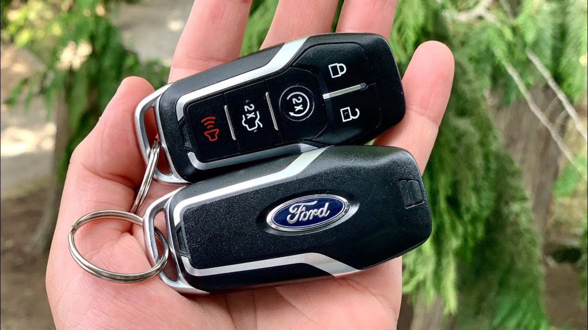 A hand holding two Ford push-to-start car keys featuring buttons for locking, unlocking, and remote starting