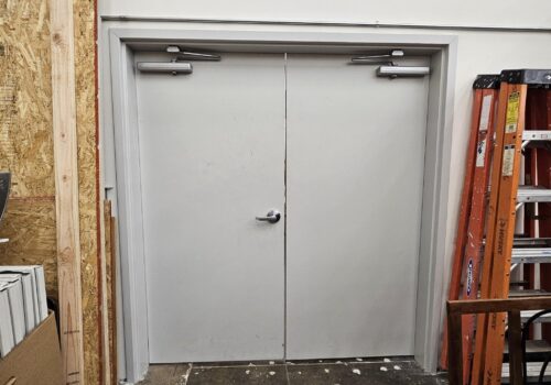 Commercial double doors with door closers, showing potential repair needs, surrounded by a construction environment with ladders and materials.