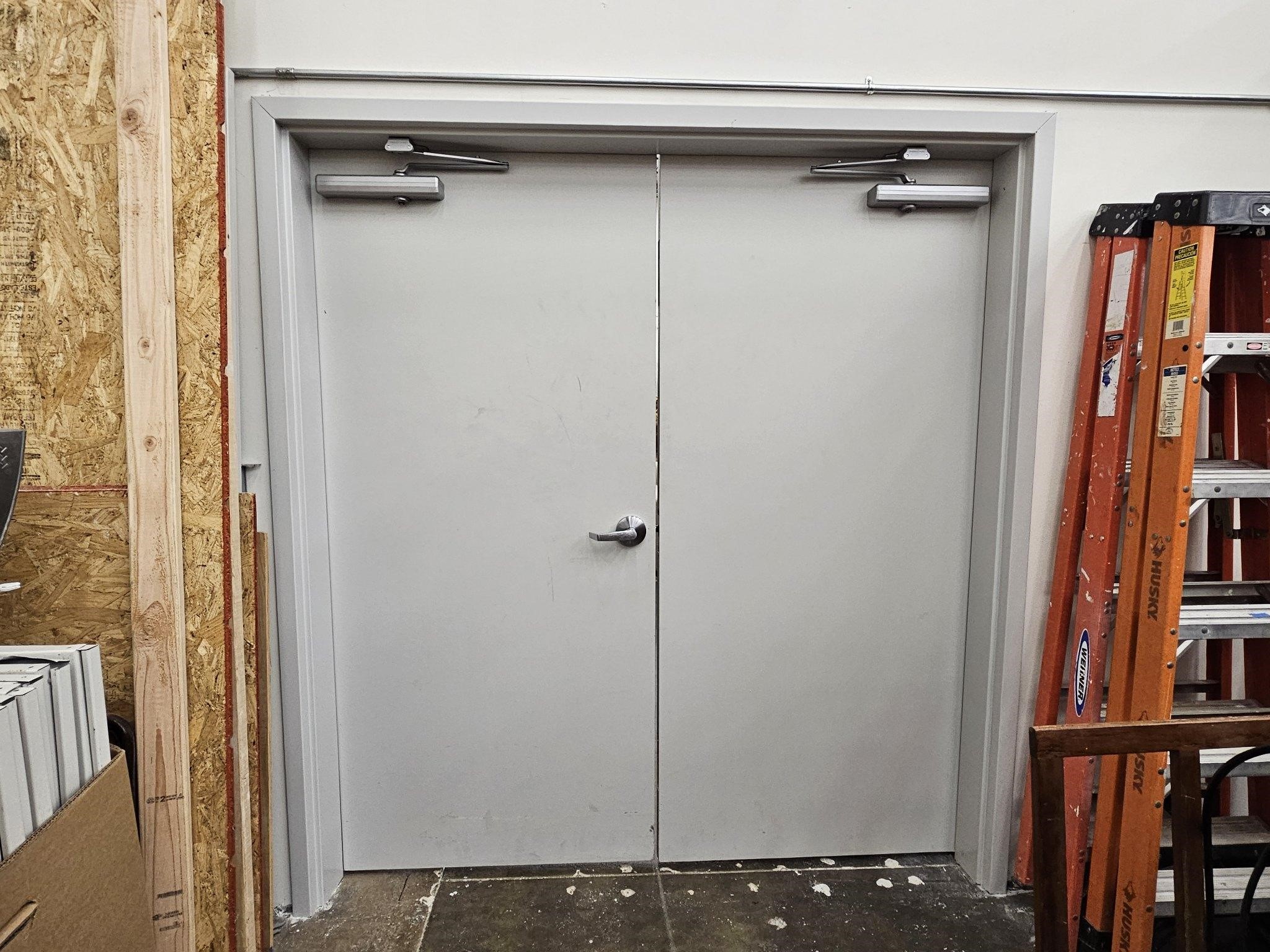 Commercial double doors with door closers, situated in a workspace with a ladder and wooden materials nearby, illustrating door functionality and repair context for MacArthur Locks & Doors services.